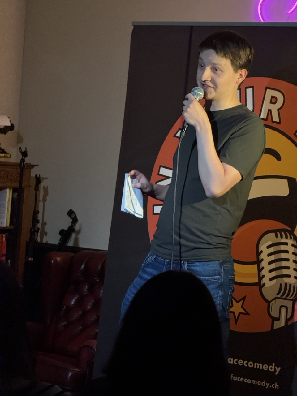 IN YOUR FACE Comedy, a comedian performing stand-up with a microphone on stage in Zürich (2026)